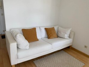 A white sofa with two brown cushions, a patterned rug, and a plain wall. - Copy of To complete (Görlitz)