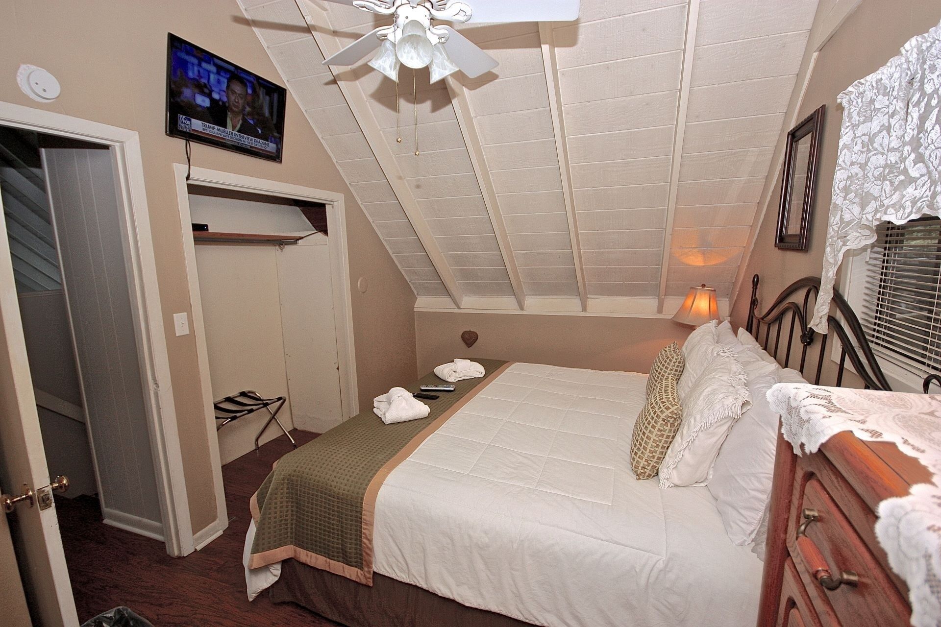 Une chambre à coucher avec un lit, un téléviseur fixé au mur, un ventilateur de plafond et une fenêtre avec des stores.
