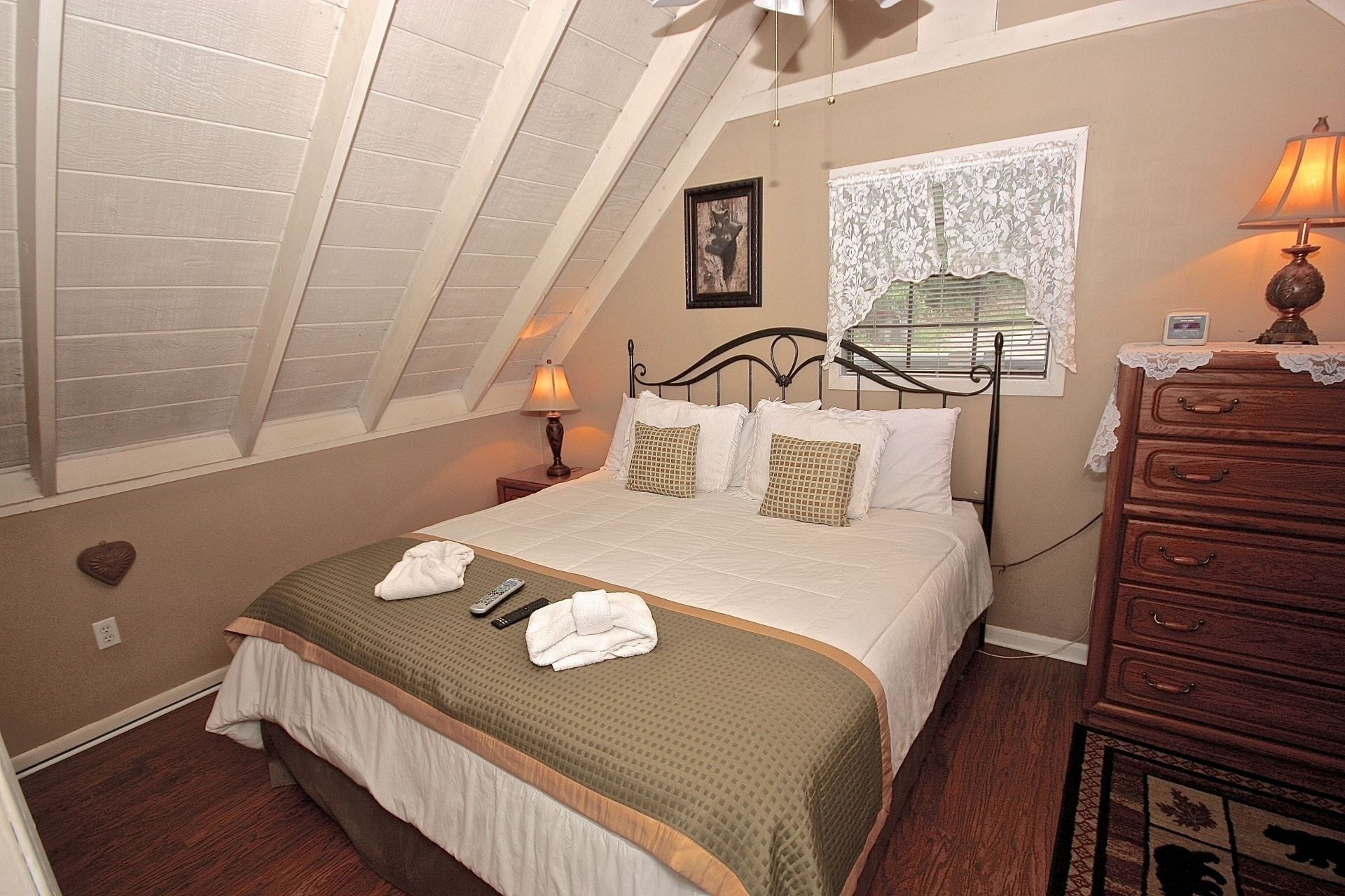 A bedroom with a sloped ceiling, a bed with a green bedspread, a dresser, a nightstand with a lamp, and a window with lace curtains.