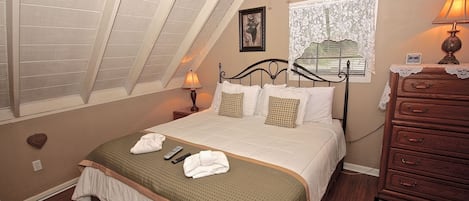 A bedroom with a sloped ceiling, a bed with a green bedspread, a dresser, a nightstand with a lamp, and a window with lace curtains.