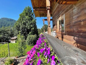 Eine Holzhütte mit Veranda, umgeben von einem Garten mit violetten Blumen, und ein Berg im Hintergrund.