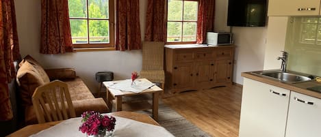 Una sala de estar acogedora con sofá, una mesa de comedor con un jarrón de flores, una zona de cocina con microondas y un televisor montado en la pared.