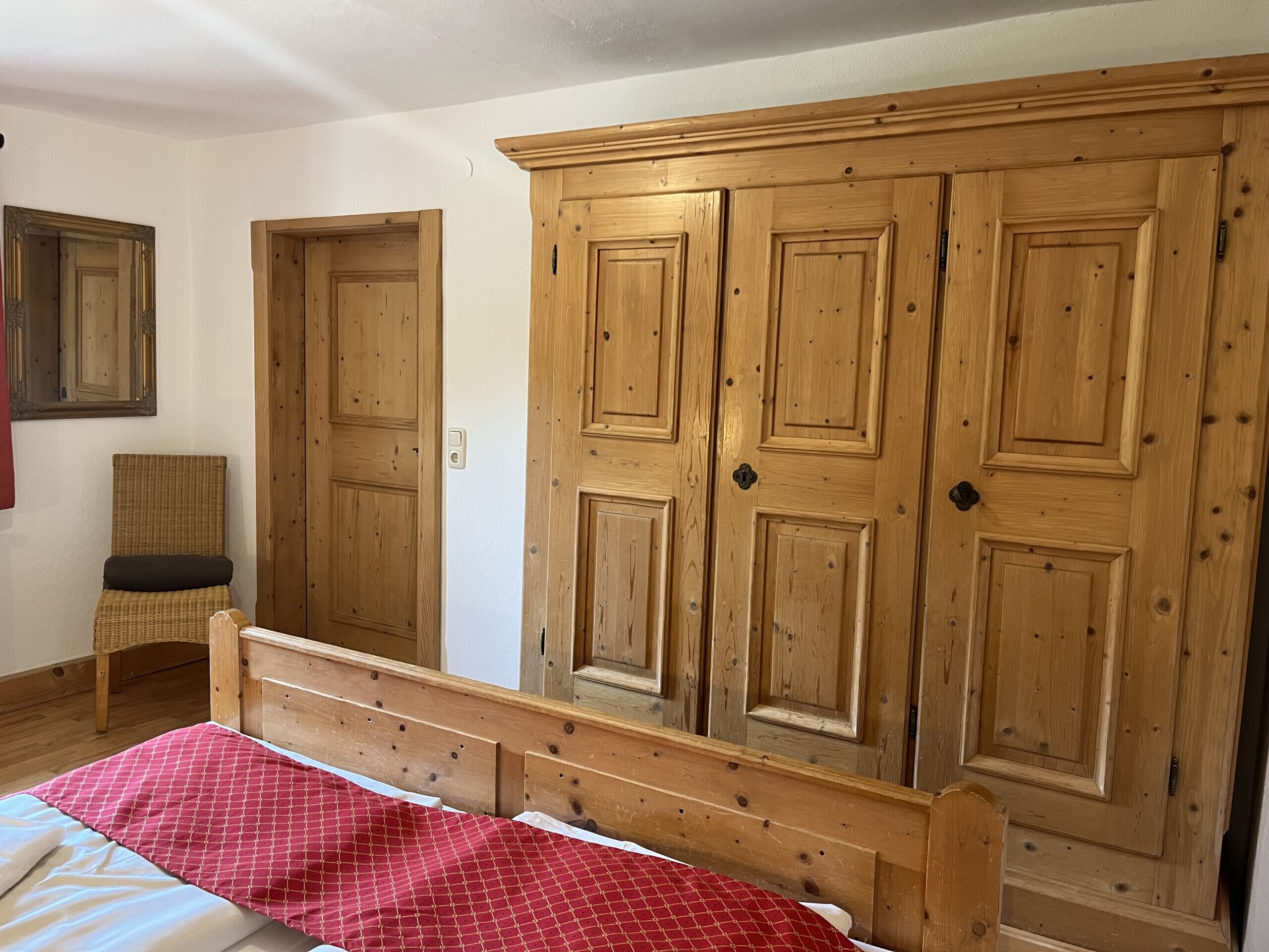 A bedroom with a wooden wardrobe, a bed with a red patterned bedspread, and a wicker chair.