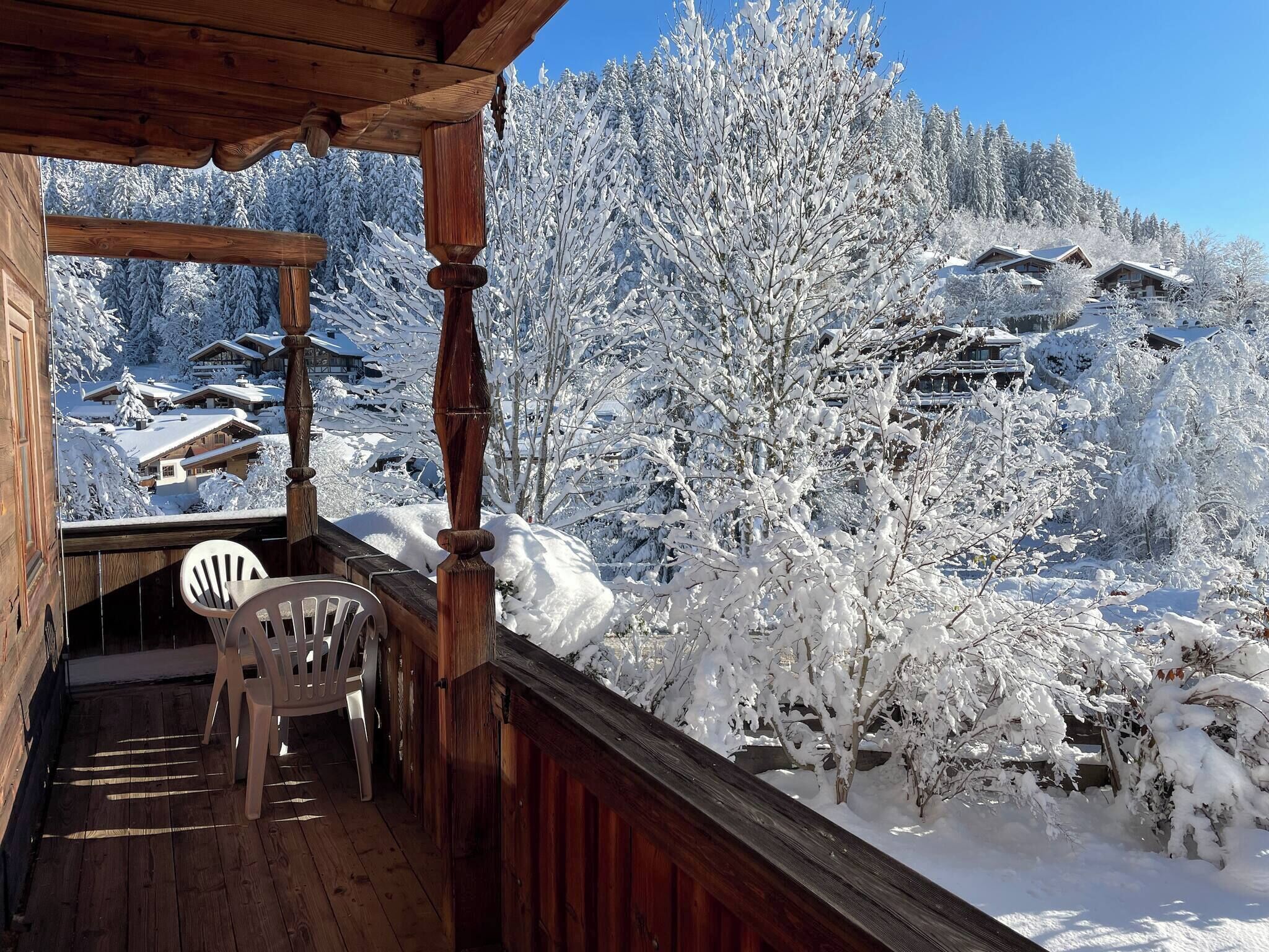 A wooden balcony with a chair, overlooking a snow-covered landscape with buildings and trees.