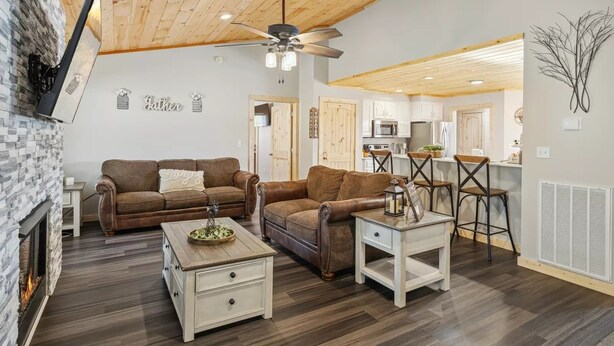 A cozy living room with a fireplace, a ceiling fan, and a kitchen area in the background. - Majestic Valley View 3 Bedroom Cabin (Sevierville)