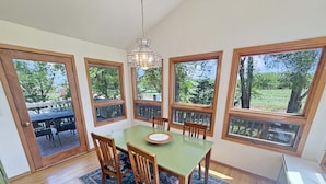 A dining area with a green table, wooden chairs, a patterned rug, and large windows with a view of trees.
