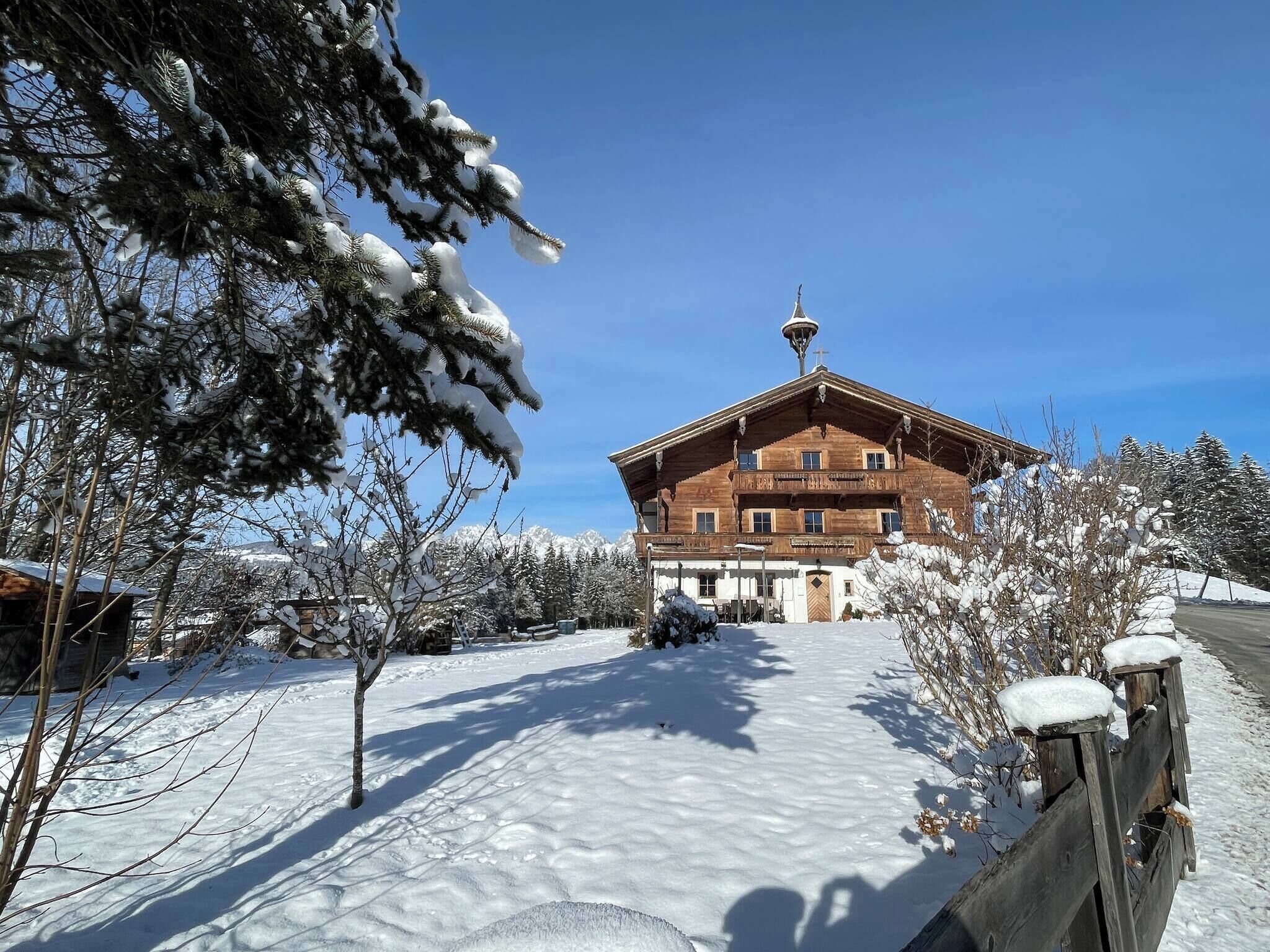 A wooden building with a sloped roof and a bell tower, surrounded by snow-covered trees and a clear sky.