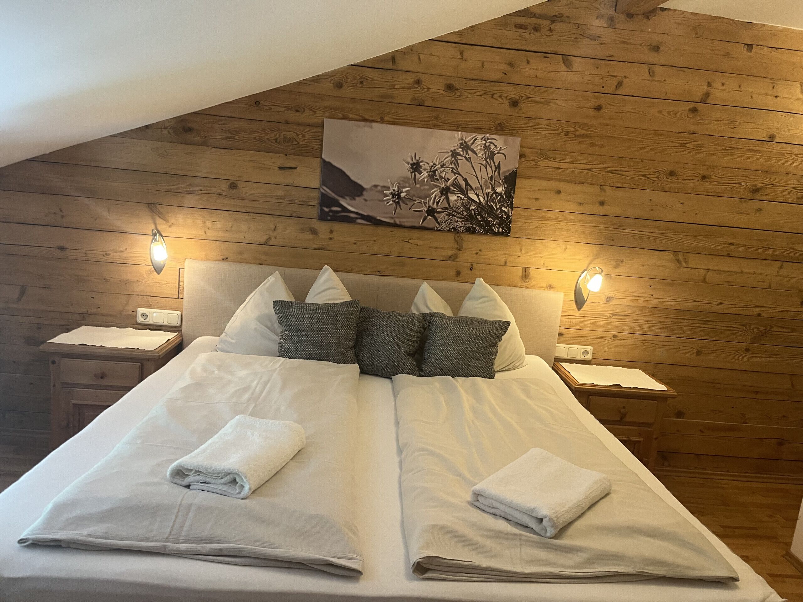 A double bed with white linens and two pillows, a wooden ceiling, and a wall-mounted picture.