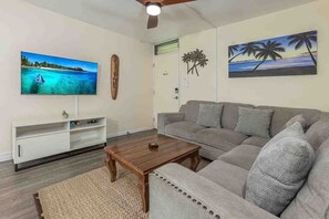 A living room with a sofa, a coffee table, a TV, and wall art. - Kalama Terrace L203 2 Bedroom Apts (Kihei)