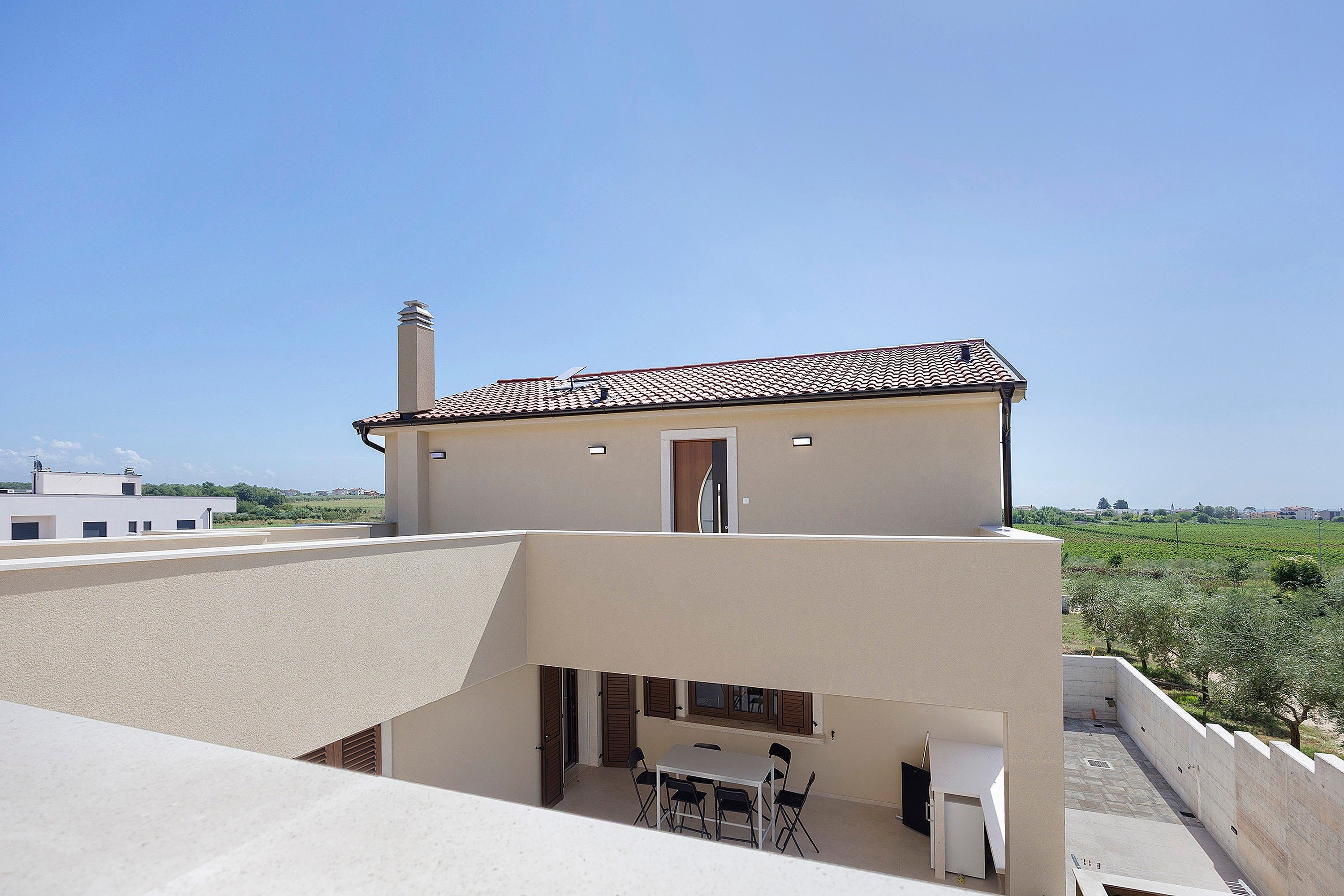 A two-story building with a flat roof, a chimney, and a balcony with a table and chairs.