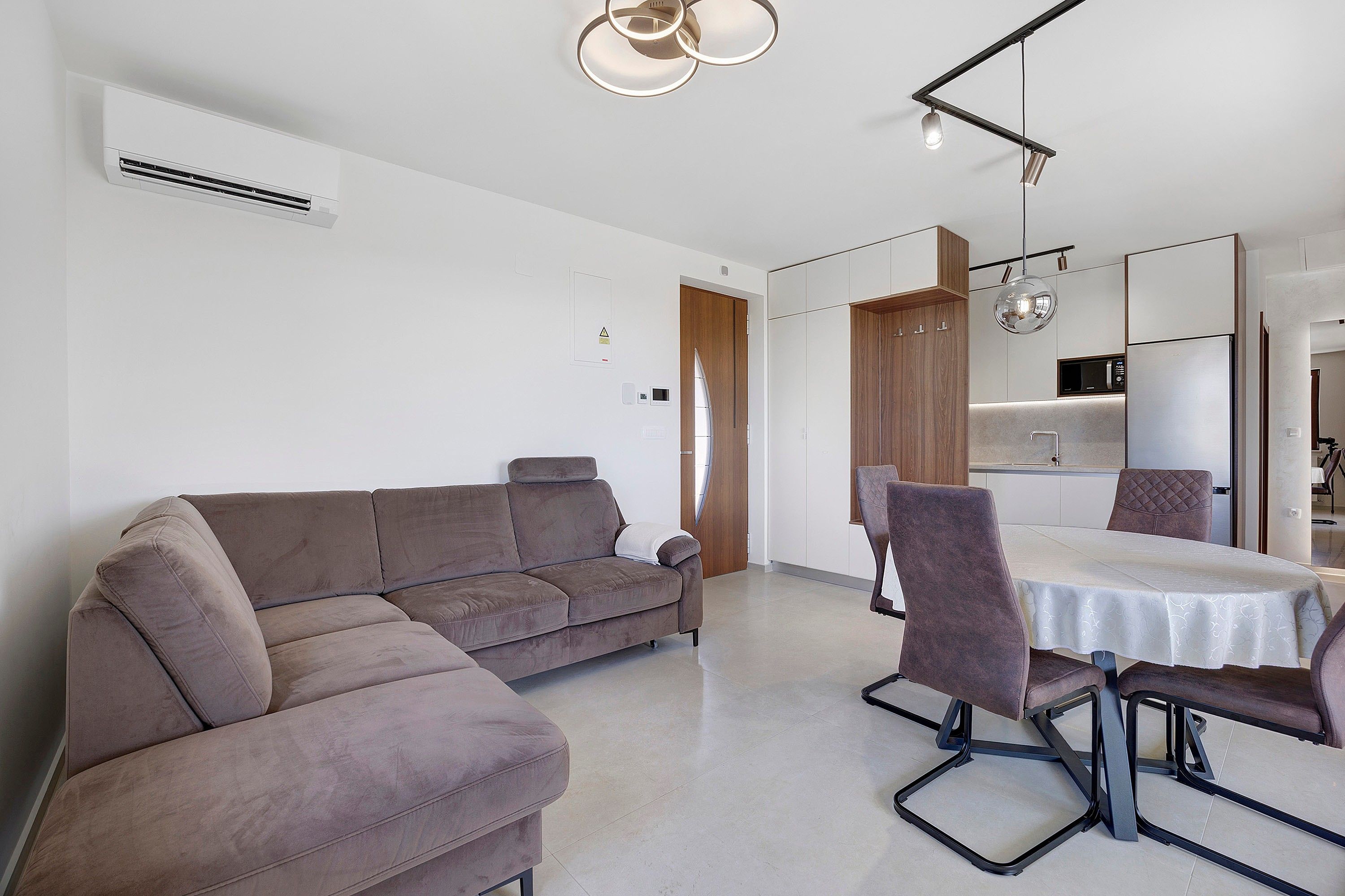 A modern living room with a sectional sofa, a dining table with chairs, and a kitchen area with a refrigerator and microwave.