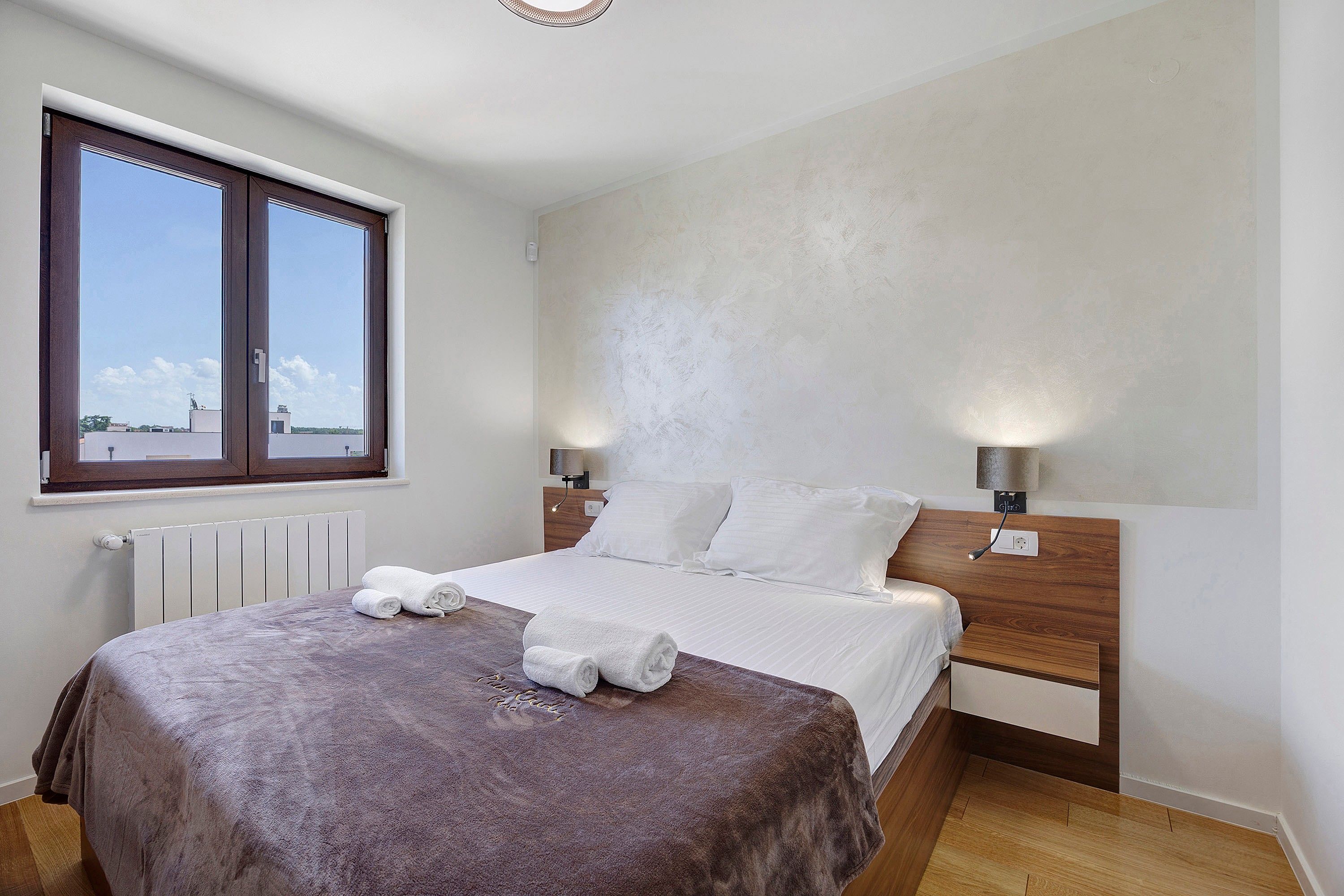 A bedroom with a bed, two bedside lamps, a window, and a radiator.