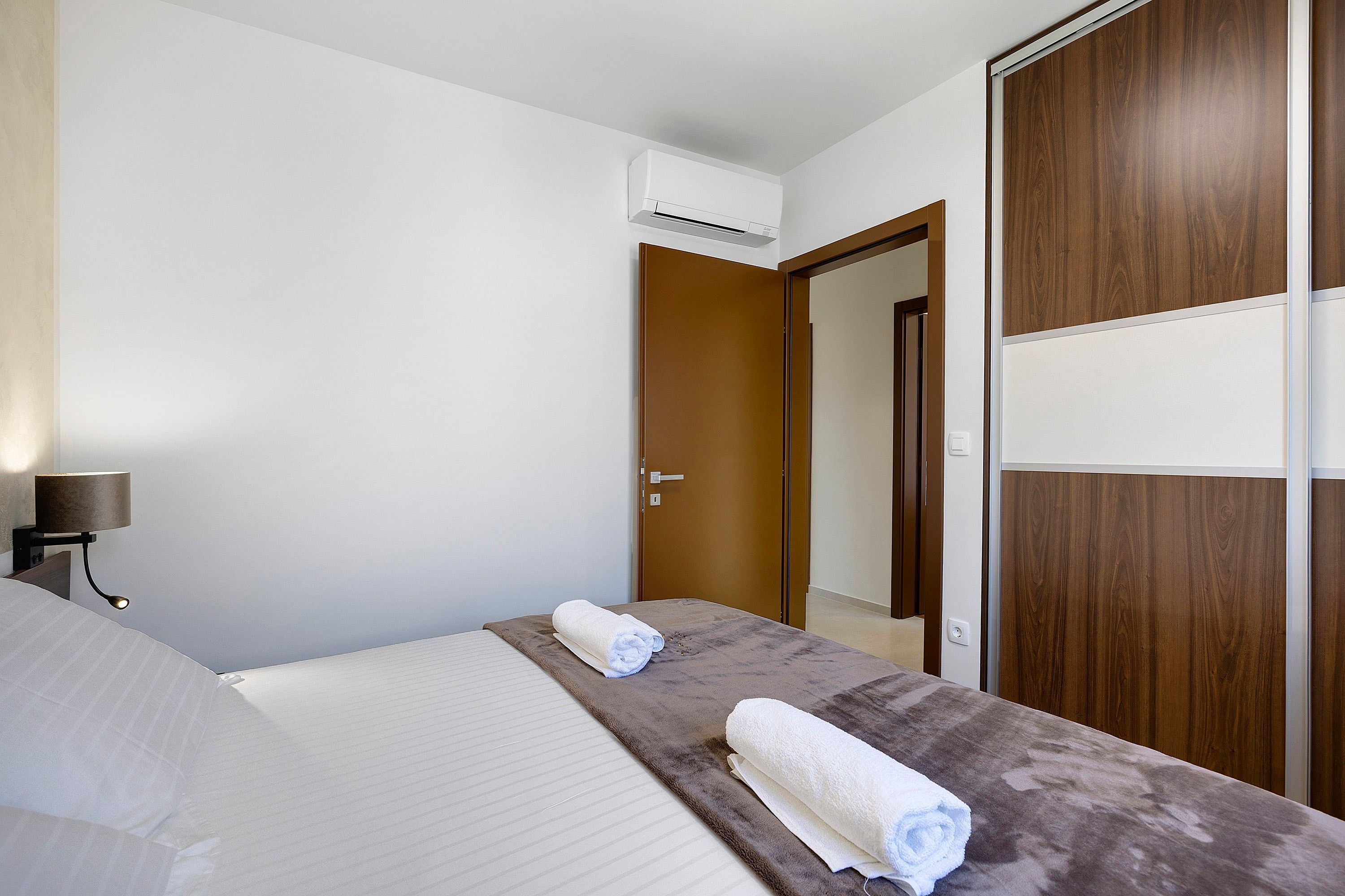 A hotel room with a bed, a nightstand, an air conditioning unit, a wooden wardrobe, and a door.