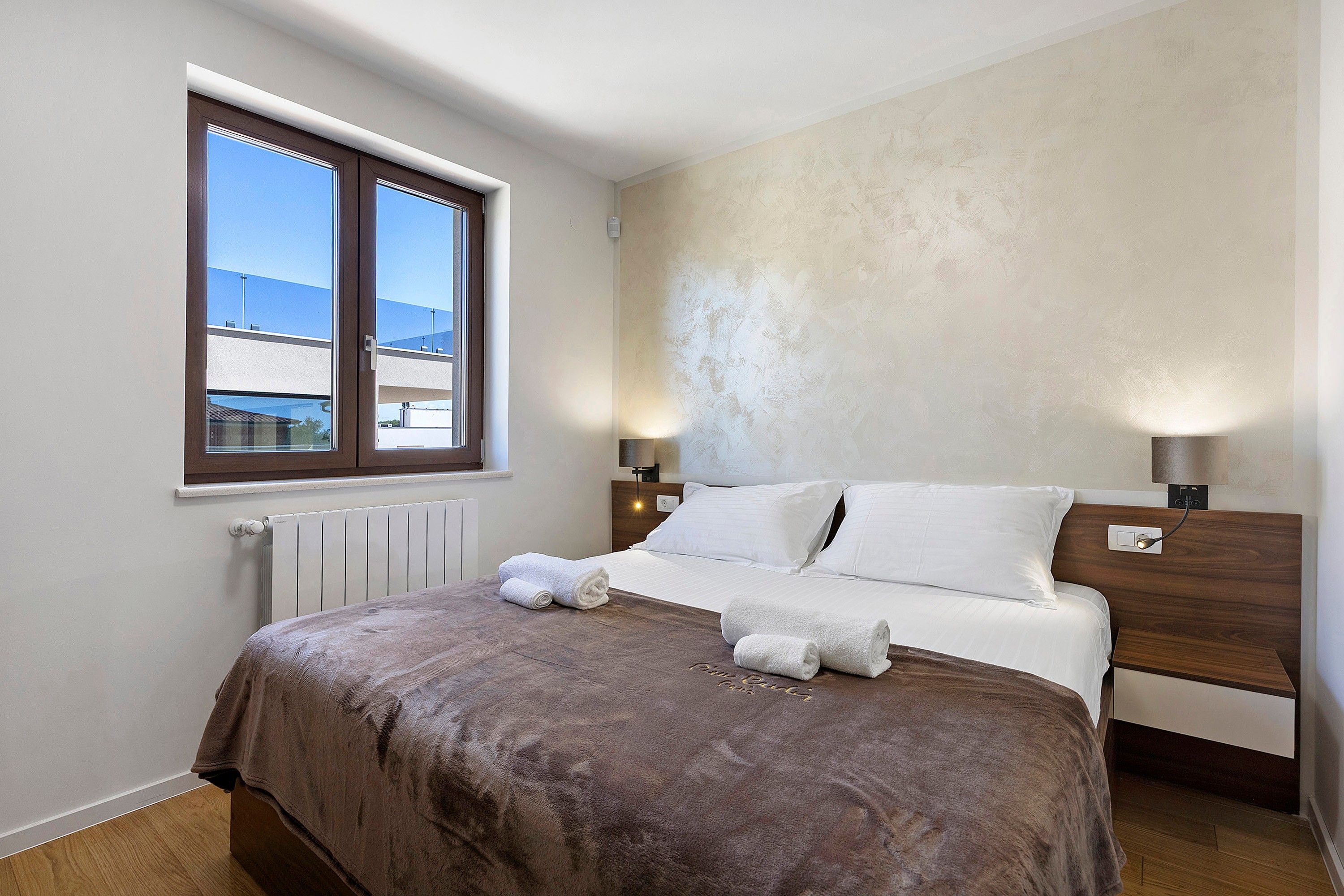 A bedroom with a bed, two bedside lamps, a window, and a radiator.