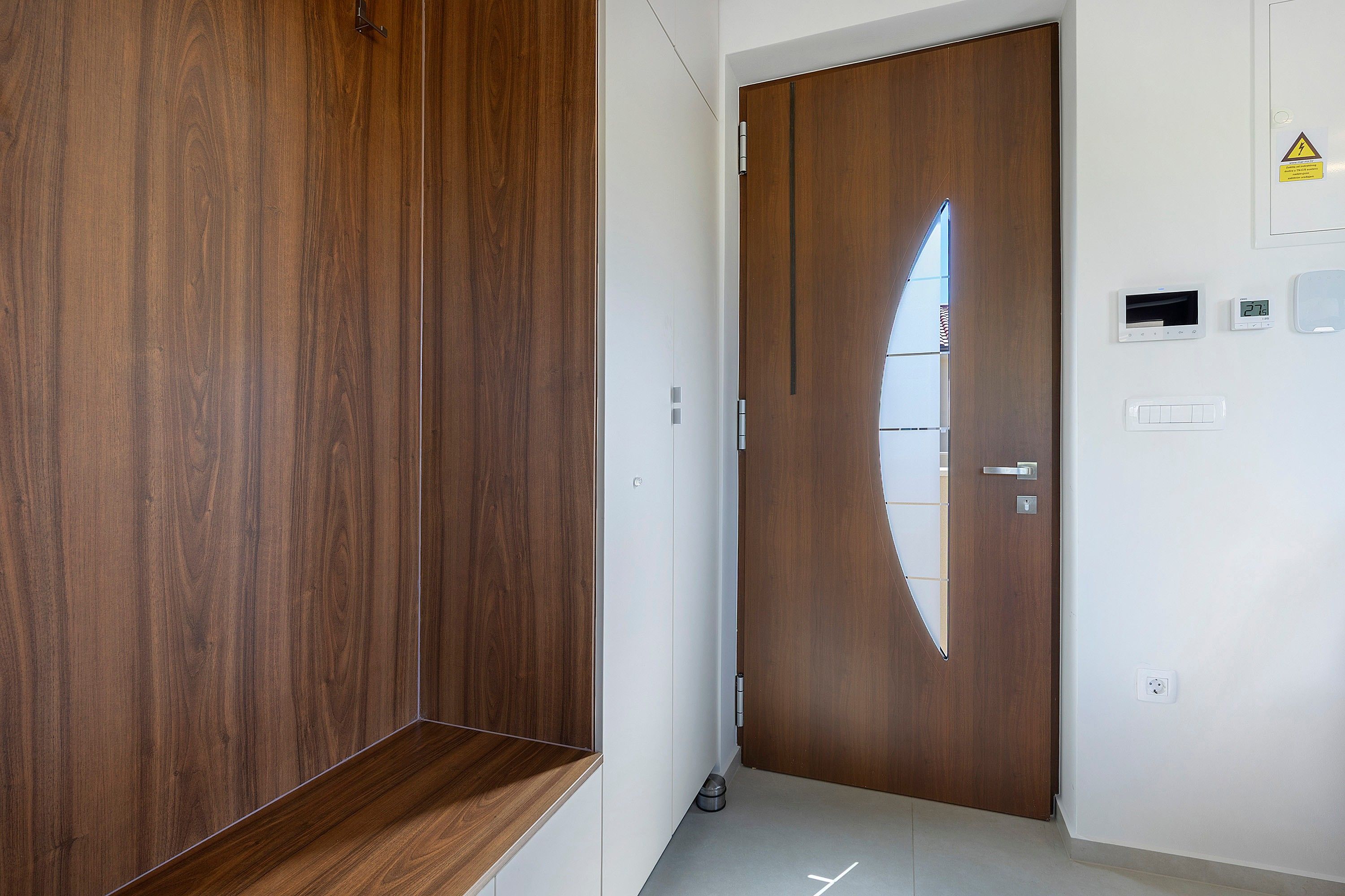 Un couloir avec une armoire en bois, une porte à surface réfléchissante et un mur muni d’un thermostat et d’une prise de courant.