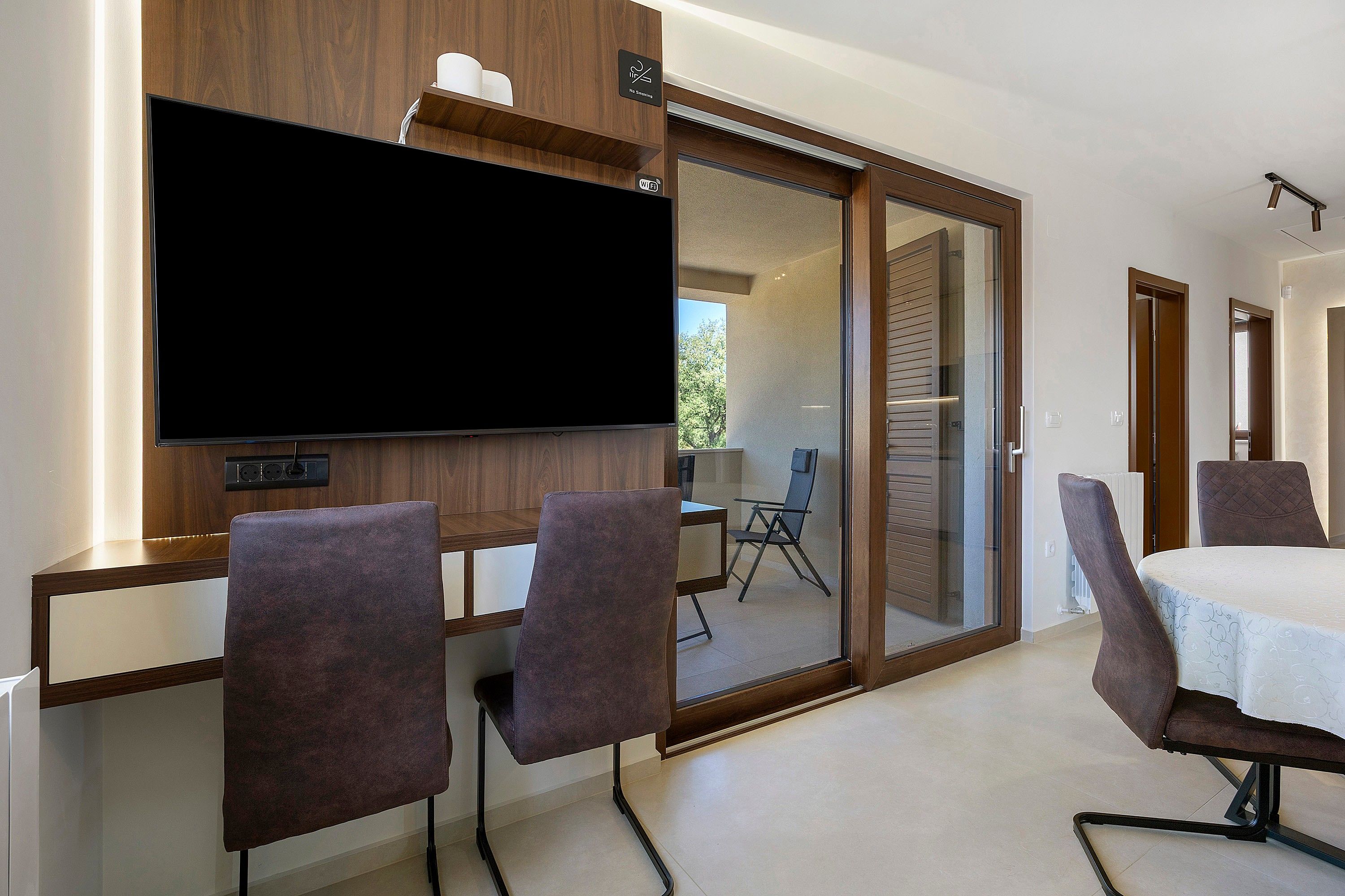 A modern room with a flat-screen TV mounted on a wooden wall, a sliding glass door leading to a balcony, and a dining area with a table and chairs.
