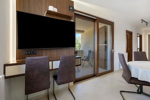 A modern room with a flat-screen TV mounted on a wooden wall, a sliding glass door leading to a balcony, and a dining area with a table and chairs.