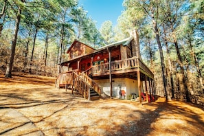 A cabin with a wooden deck, a staircase leading to the entrance, and a surrounding forest. - Nestled in Nature: Pet-Friendly Family Cabin w/ Hot Tub, Arcade & Playground (Broken Bow)
