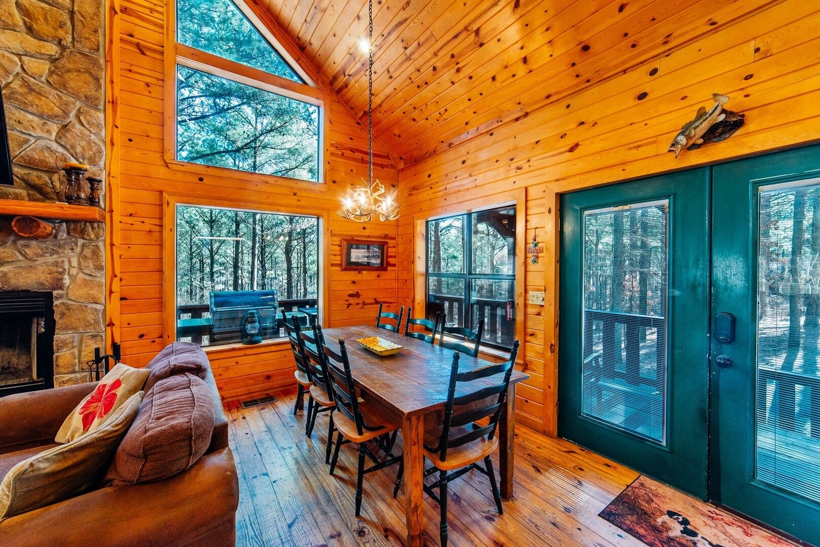 Een chaletinterieur met een stenen openhaard, een houten eettafel en grote ramen met uitzicht op het bos.