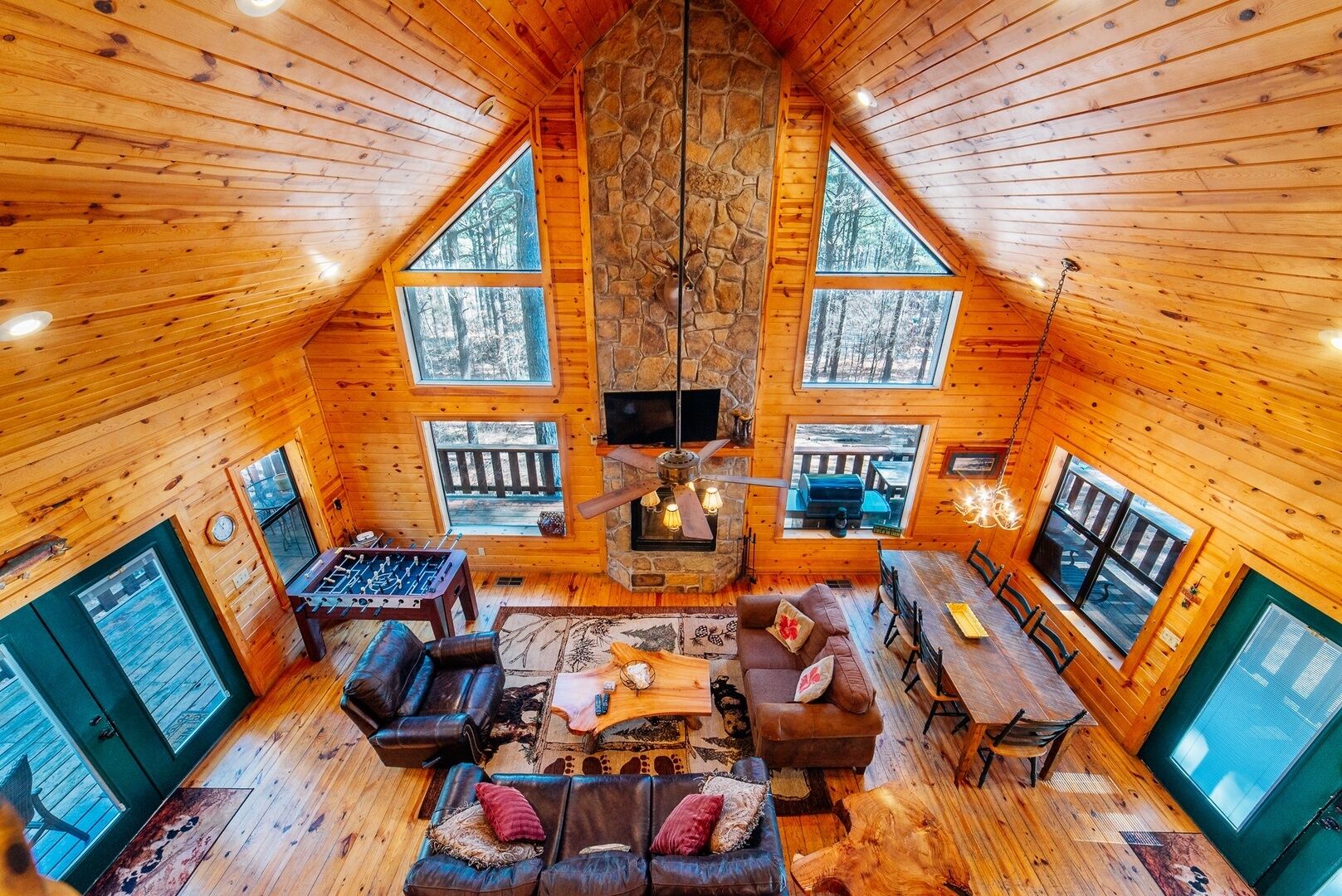 A cabin interior with a high ceiling, wooden walls, a fireplace, a dining area, and large windows.