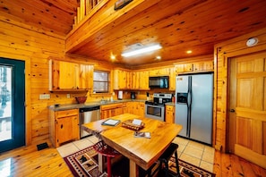 A wooden cabin interior with a kitchen featuring a central island, stainless steel appliances, and a window with blinds. - Nestled in Nature: Pet-Friendly Family Cabin w/ Hot Tub, Arcade & Playground (Broken Bow)