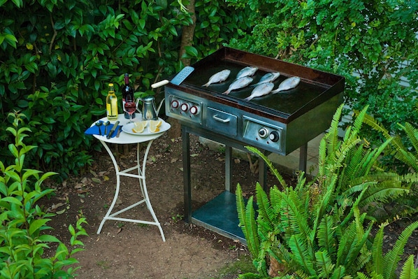 An outdoor grill with fish cooking, a table with a bottle of wine, and a glass of red wine.