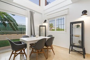 A modern dining area with a white table and chairs, a glass cabinet, and a view of a garden through large windows.