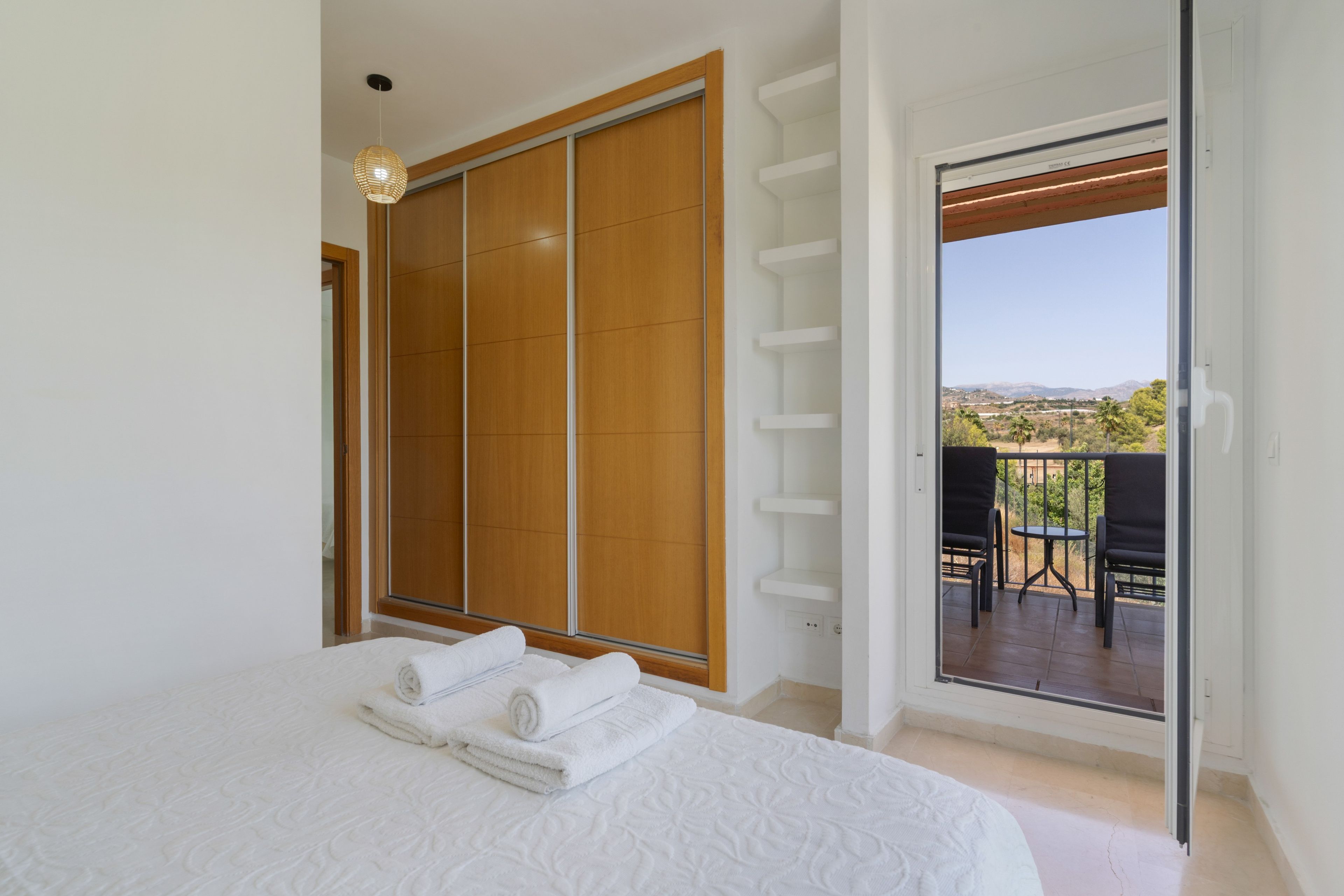 A bedroom with a bed, a wooden wardrobe, and a balcony with a table and chairs.