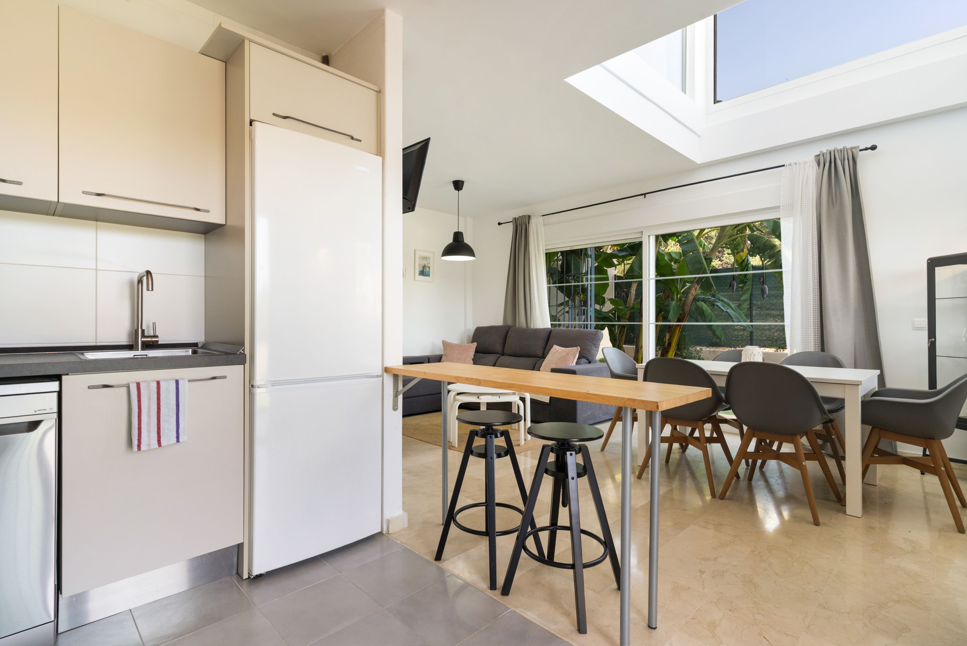 Eine moderne Küche mit einem eingebauten Kühlschrank, ein Essbereich mit einem Holztisch und Stühlen sowie ein großes Fenster mit Vorhängen.