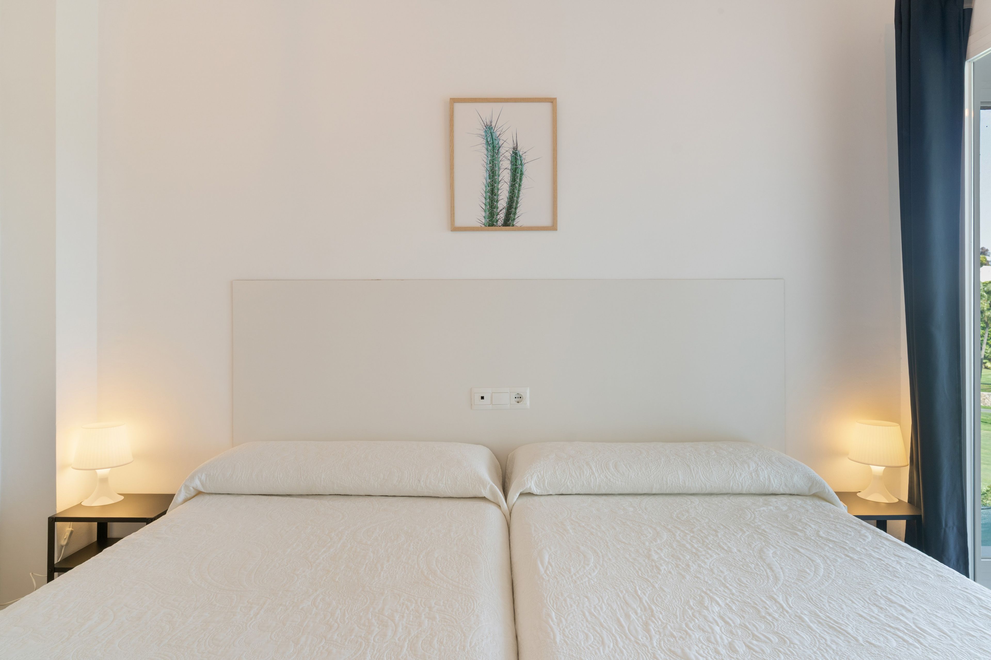 Een tweepersoonsbed met hoofdeinde, twee bedlampen, een stopcontact en een lijst met een cactusschilderij aan de muur.