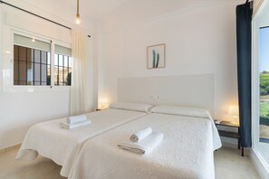 A hotel room with two beds, a window with curtains, a wall-mounted picture, and a bedside table with a lamp.