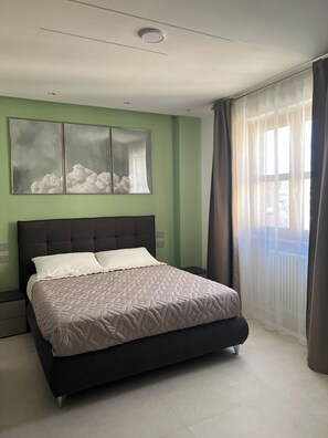 Un dormitorio con cama, mesita de noche, lámpara de pared y ventana con cortinas.