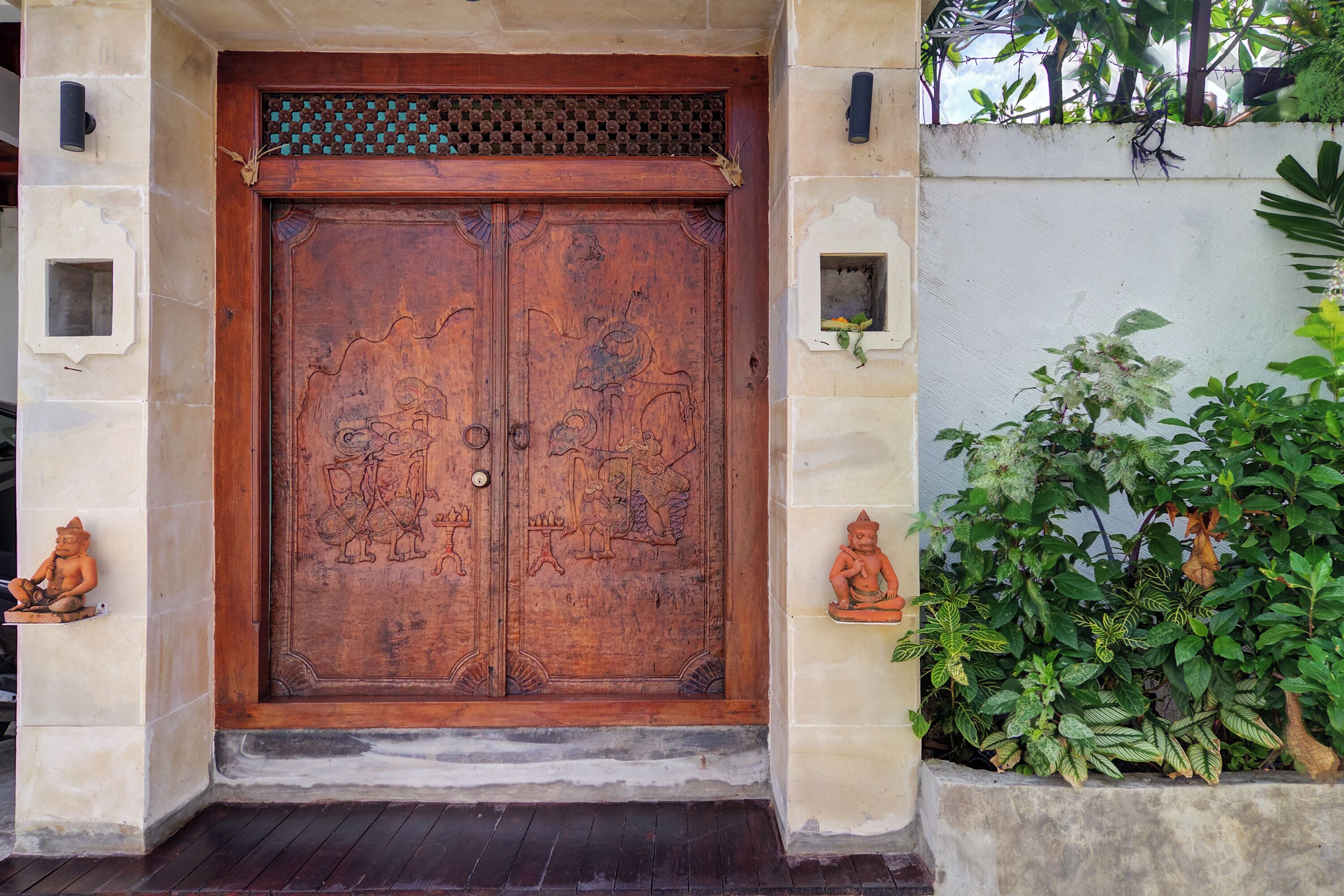 Una puerta de madera grande con intrincados relieves, flanqueada por dos pequeñas estatuas y una planta en maceta.
