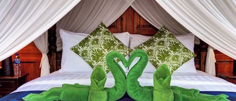 Una cama con sábanas blancas, dos almohadas verdes con estampado de hojas y una toalla verde formando un corazón.
