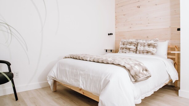 Ein schlichter Schlafzimmerraum mit einem hölzernen Kopfteil, einem weißen Bett und einem schwarzen Stuhl.