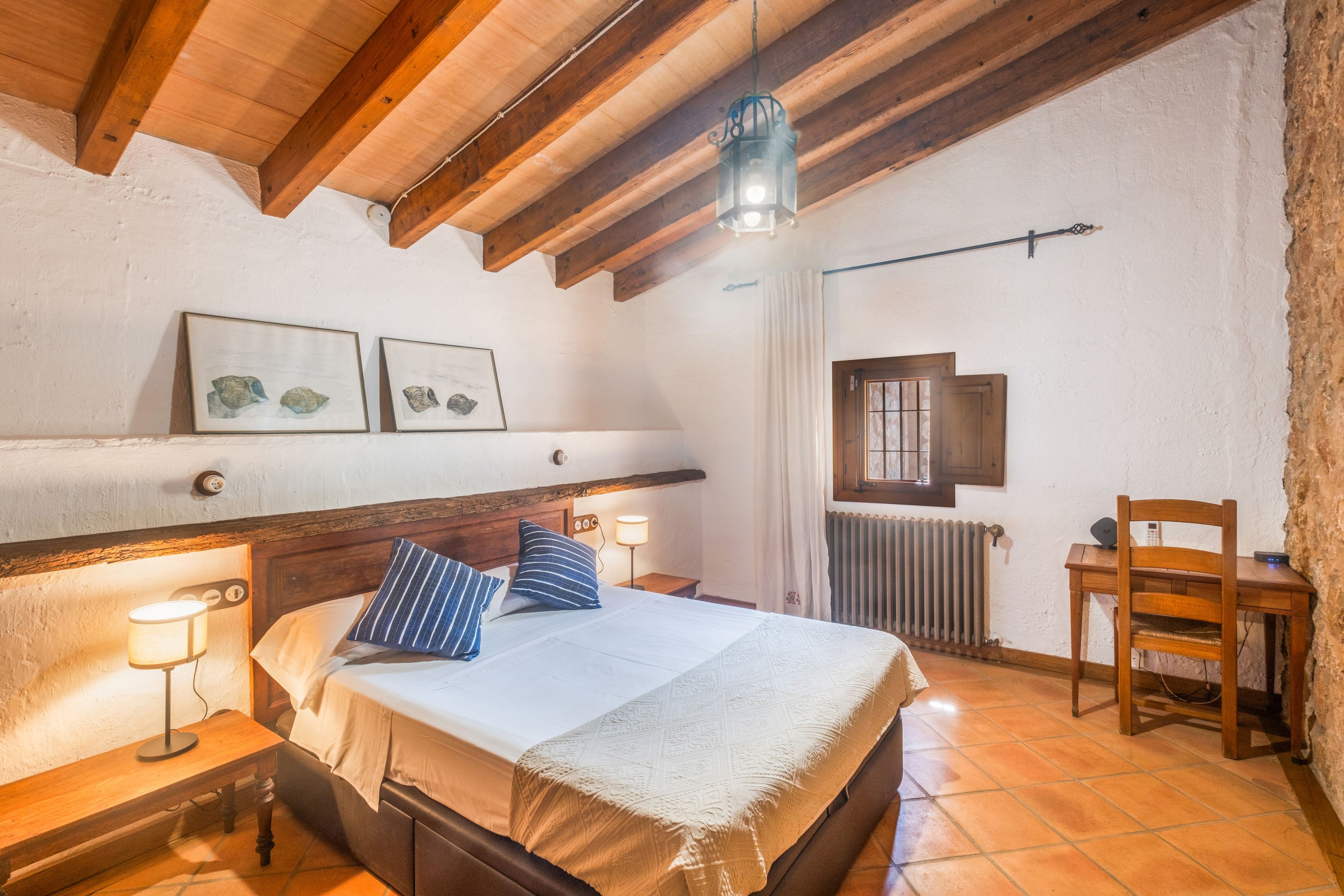 Un dormitorio con techo de madera, una cama con colcha blanca y almohadas azules, una mesita de noche con lámpara, una silla y un radiador.