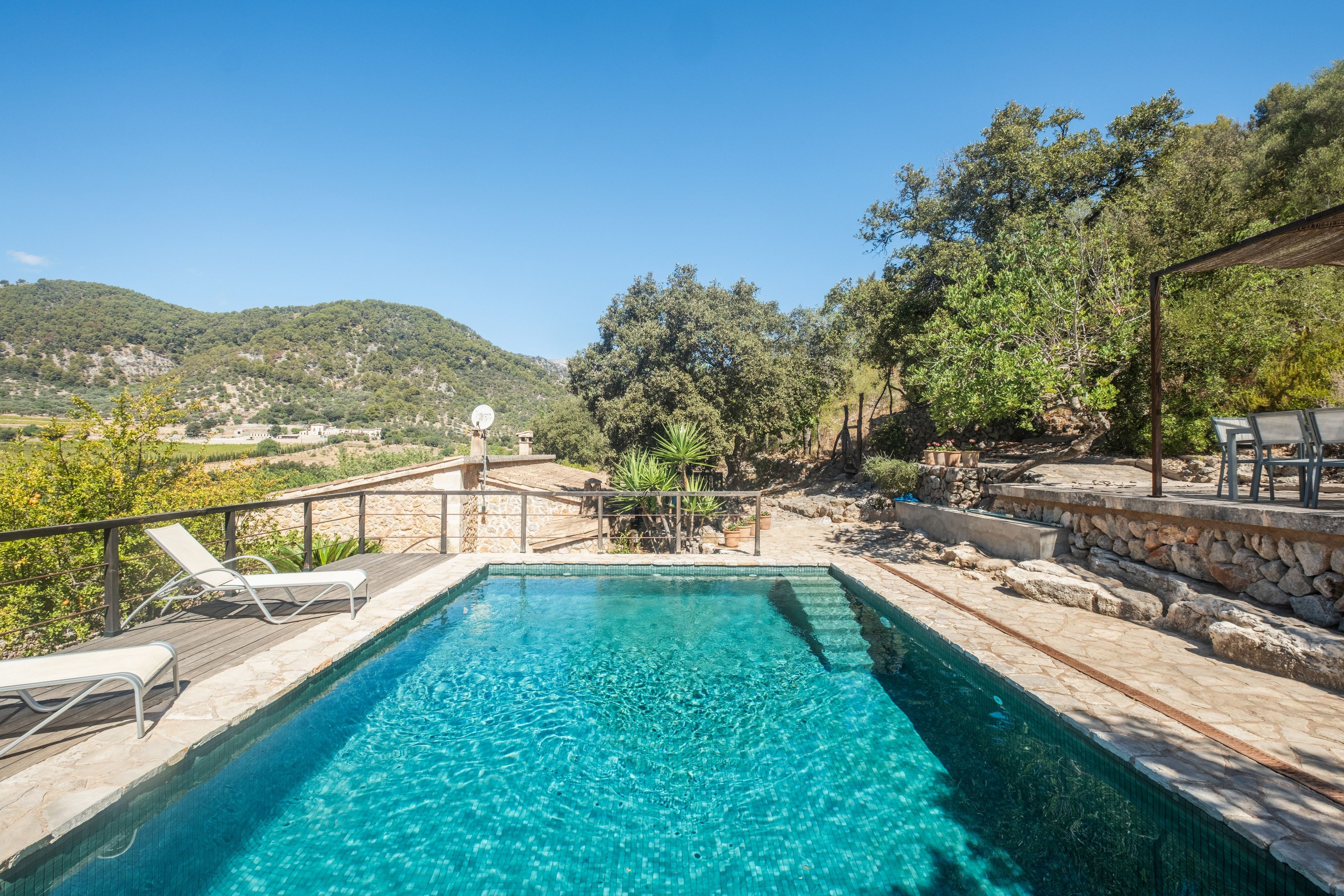 Una piscina azul cristalina con un muelle de madera y sillas de descanso, rodeada de muros de piedra y vegetación.