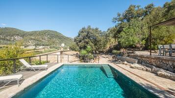 Una piscina azul cristalina con un muelle de madera y sillas de descanso, rodeada de muros de piedra y vegetación.