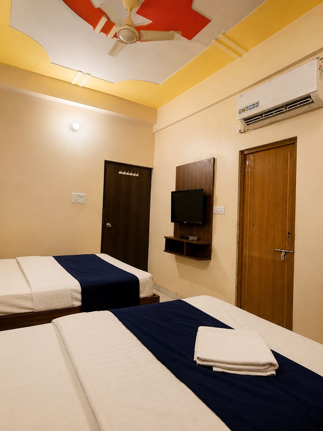 A hotel room with two beds, a ceiling fan, an air conditioning unit, a television, and a wooden door.
