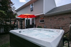 Una bañera de hidromasaje en un patio trasero con una sombrilla roja, una casa de ladrillo y una terraza de madera.