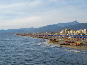 Una playa costera con numerosos paraguas y barcos, un cielo despejado y montañas al fondo.
