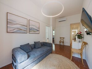 A modern living room with a grey sofa, two abstract paintings, a round ceiling light, and a dining area with a wooden chair and a potted plant. - Beautiful  villa for 5 people with WIFI, A/C, TV and patio (Viareggio)