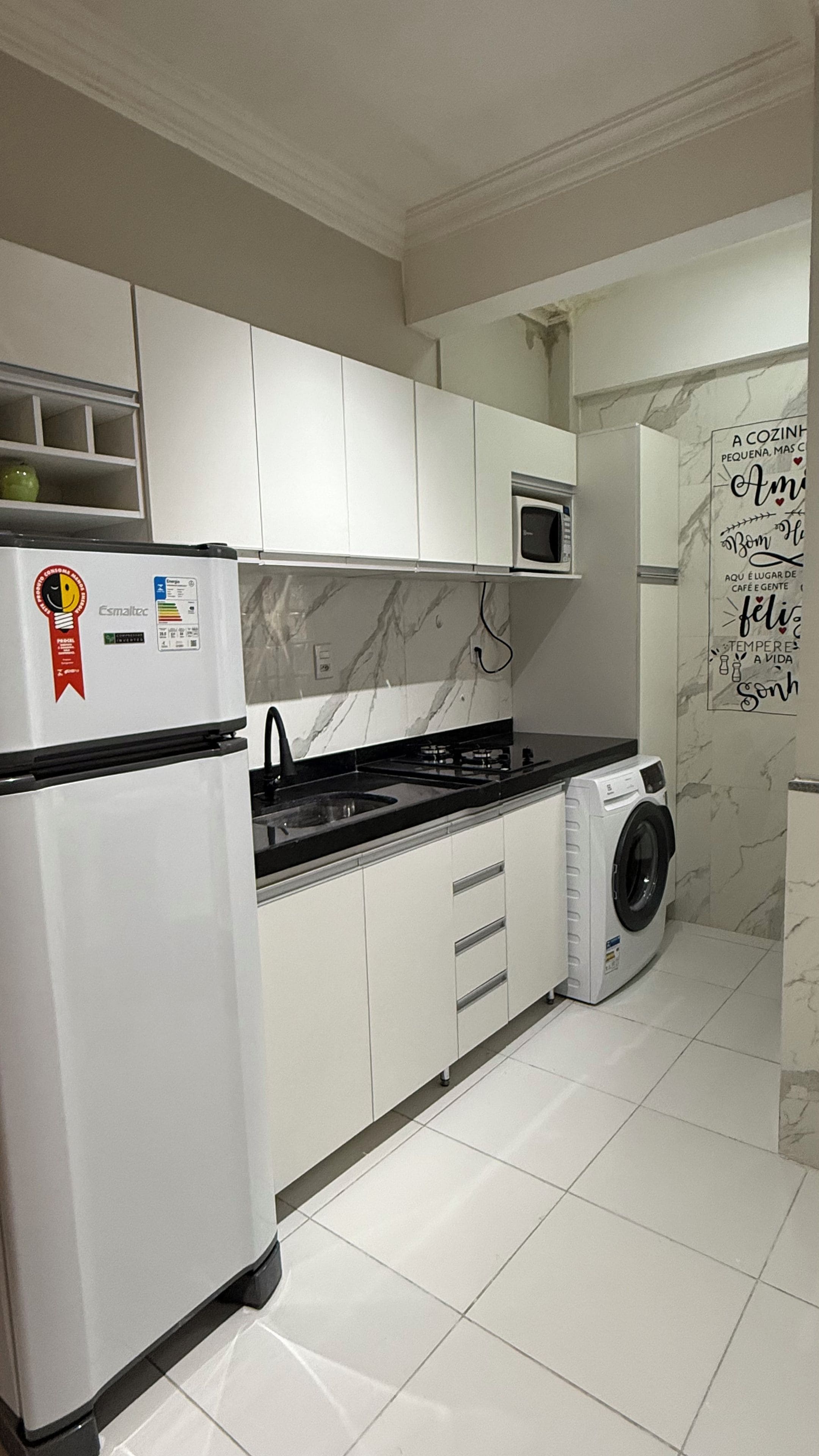 Una cocina con armarios blancos, un microondas integrado, una lavadora y un refrigerador con un adhesivo.
