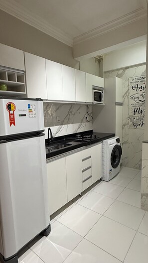 A kitchen with white cabinets, a built-in microwave, a washing machine, and a refrigerator with a sticker. - 105 AP 2 Qtos Prático e Funcional (Belém)