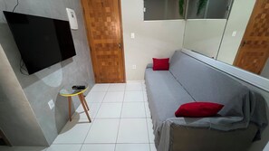 A small, compact room with a flat-screen TV mounted on the wall, a small round table with a remote control, a grey sofa with a red pillow, and a wooden door. - 105 AP 2 Qtos Prático e Funcional (Belém)