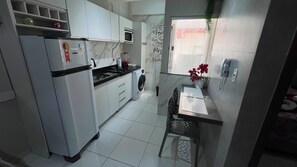 A compact kitchen with a refrigerator, microwave, and a washing machine. There is a dining area with a table and chairs. - 105 AP 2 Qtos Prático e Funcional (Belém)