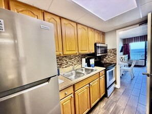 A compact kitchen with a refrigerator, microwave, and stove. There is a sink and wooden cabinets. A balcony with a view is visible through an open door. - Oceanfront 1BR Sleeps 4-6 Myrtle Beach 421 (Myrtle Beach)