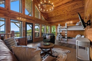 A cabin interior with a wooden ceiling, large windows, a sofa, a table, and a bunk bed. - MTN Views|2 King Suites|Private|HotTub|Game Room|+ (Gatlinburg)