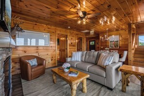 Interior de uma cabana de tronco com lareira, um sofá, uma mesa de centro e uma mesa de jantar.