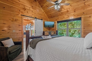 A wooden cabin bedroom with a bed, a television, and a window with a view of greenery. - MTN Views|2 King Suites|Private|HotTub|Game Room|+ (Gatlinburg)